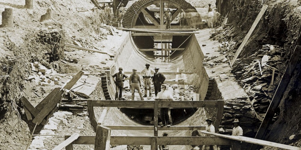 Historic photo dated April 30, 1901 showing the Wingohocking sewer under construction on Courtland St. From the City Archives of Philadelphia. Working men stand in various places in and around an incomplete tunnel made of bricks and stone and held in a tubular shape by wooden support beams. A horse or mule hitched to a small cart and several other men stand on a makeshift bridge across the ditch in which the sewer tunnel lies. A forest of hardwood tree trunks is visible in the background and a few stumps of cut trees are visible on the banks of the ditch.